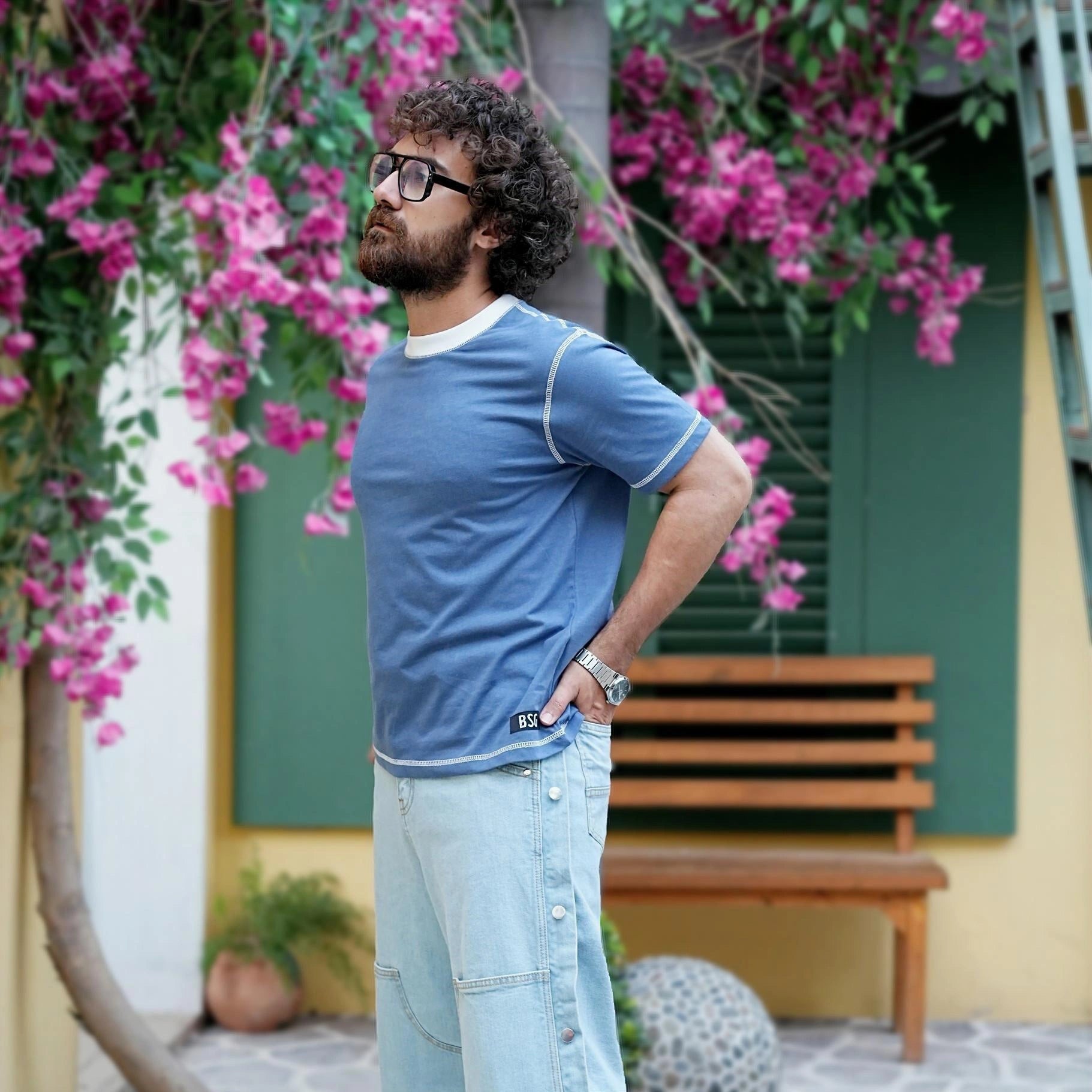 Man in a blue shirt standing in front of a building with pink flowers and a bench.