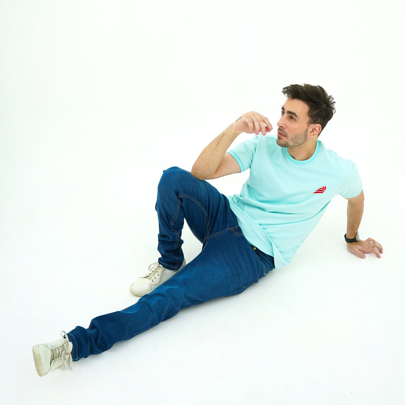 Man sitting on a white floor wearing BSG light blue t-shirt and dark blue jeans.