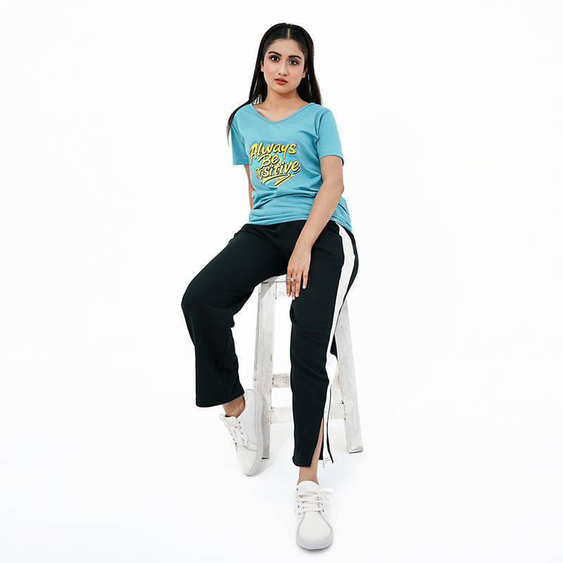 Woman sitting on a stool wearing a blue t-shirt with yellow text and black pants with a white stripe.