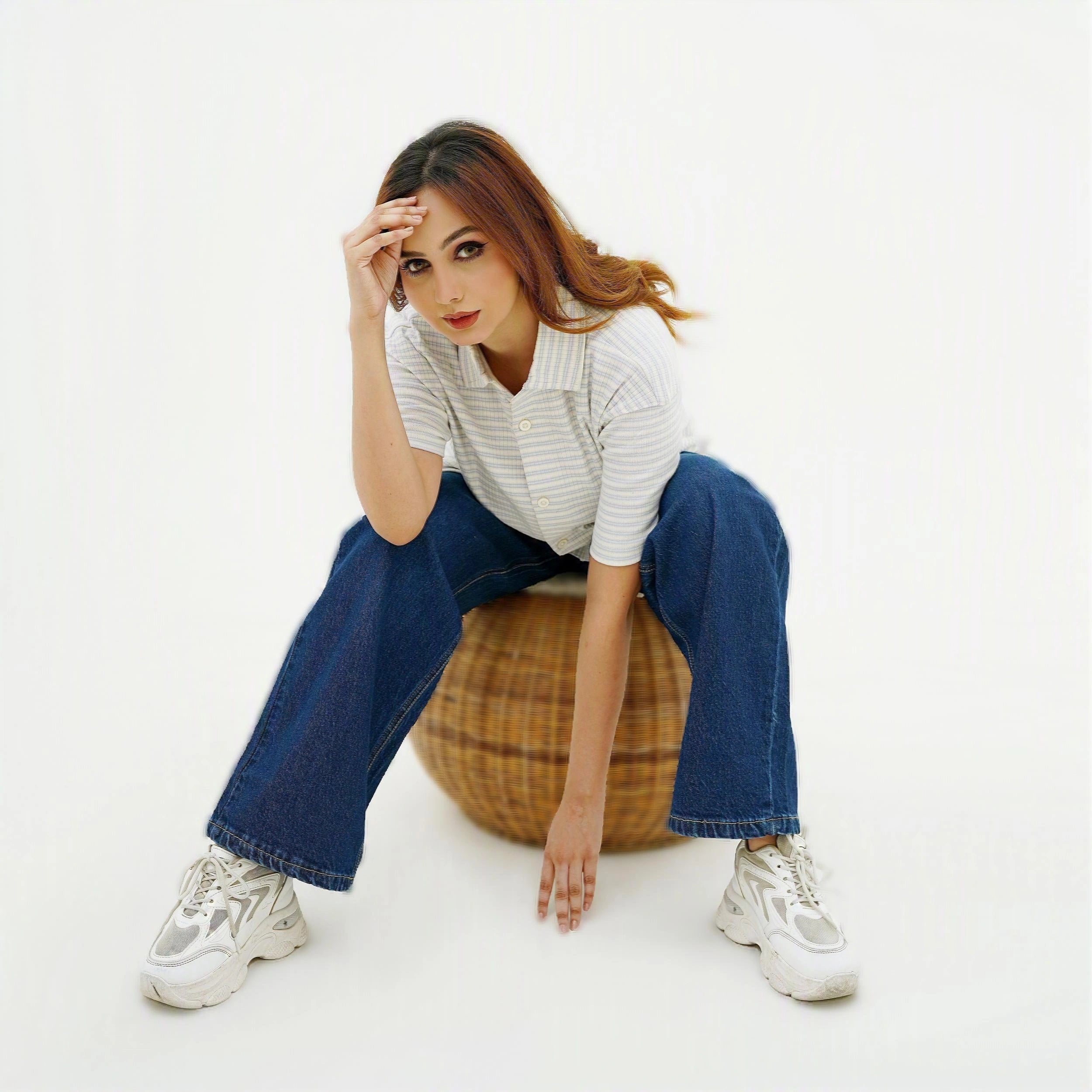 Woman sitting on a woven ball wearing a striped shirt, jeans, and sneakers on a white background