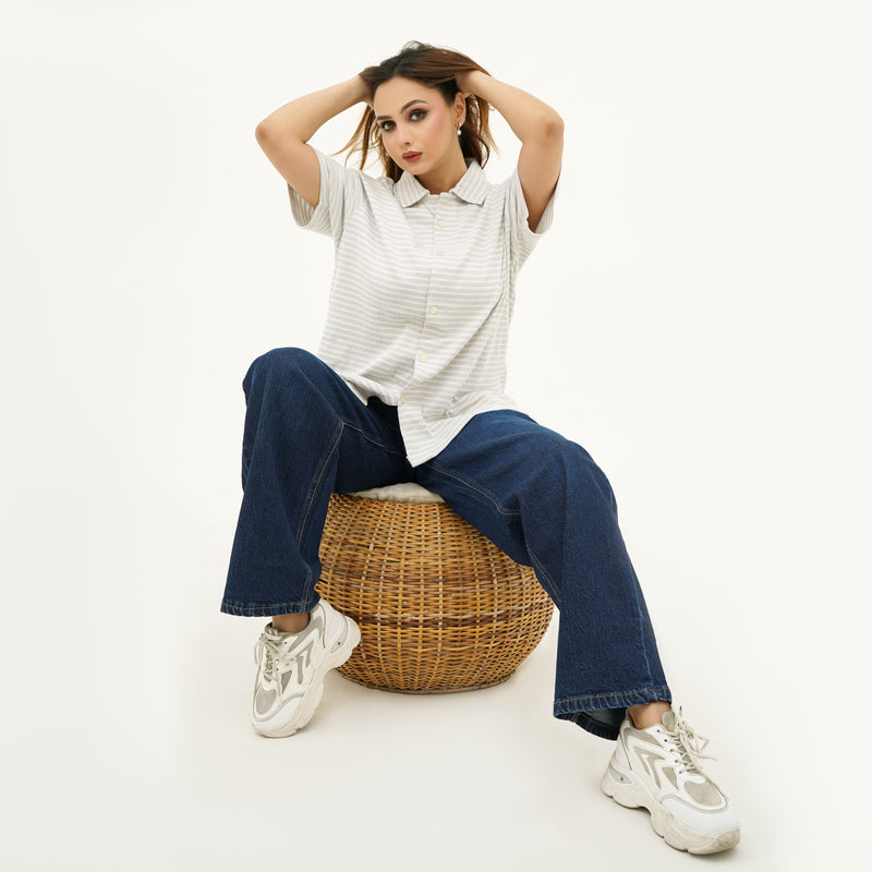 Woman sitting on a wicker stool wearing BSG white shirt, blue jeans, and white sneakers.