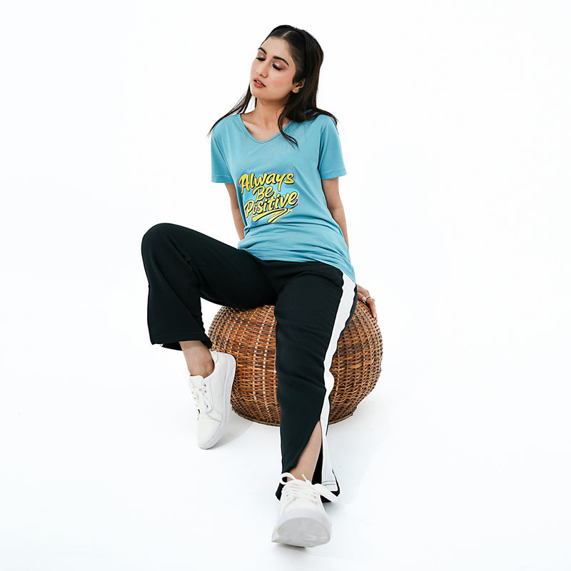 Woman sitting on a woven stool wearing a blue t-shirt with text and black pants.